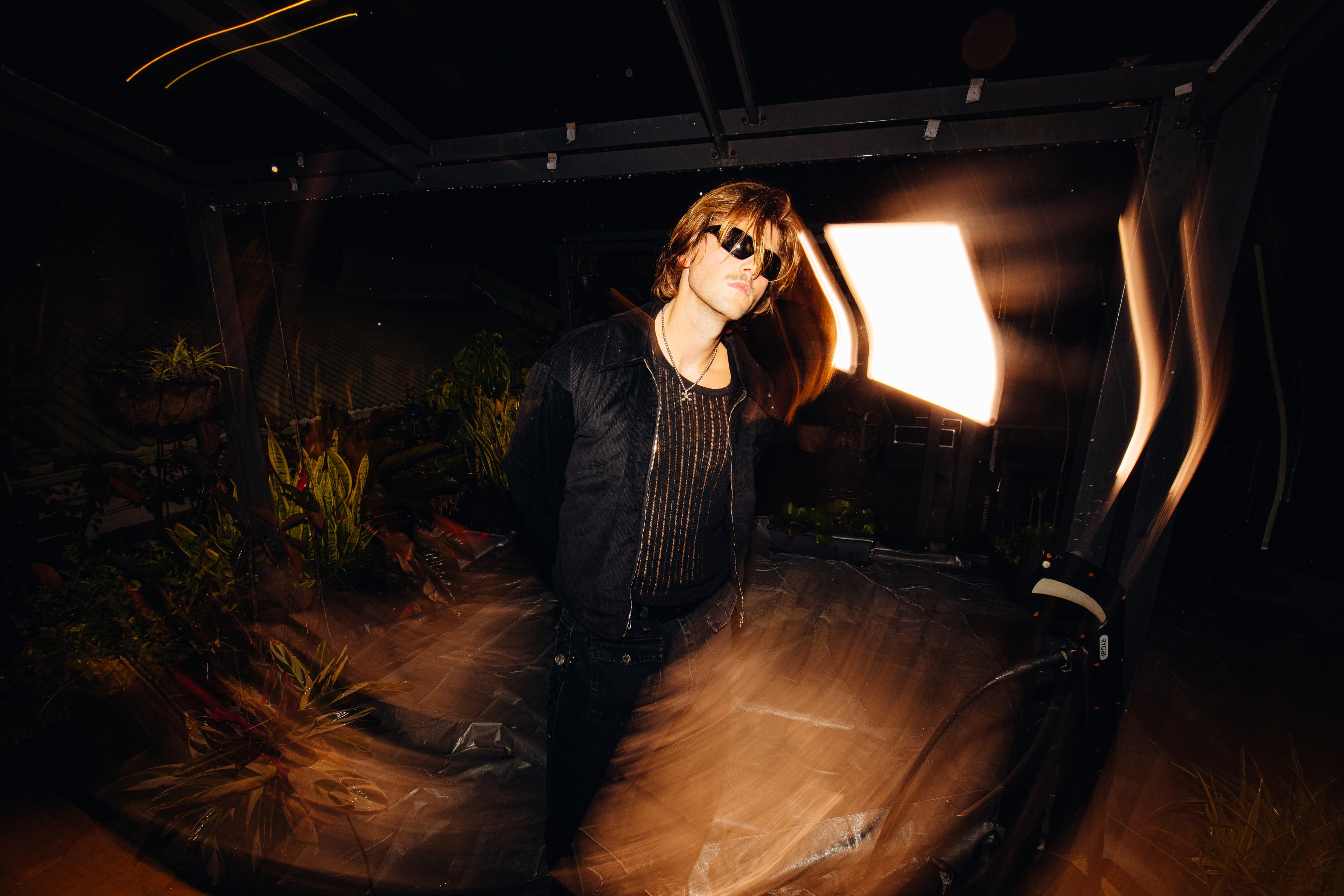 Ruel in sunglasses and a black jacket, lit by a studio light with long-exposure light trails, at Mood on the Roof in Sydney