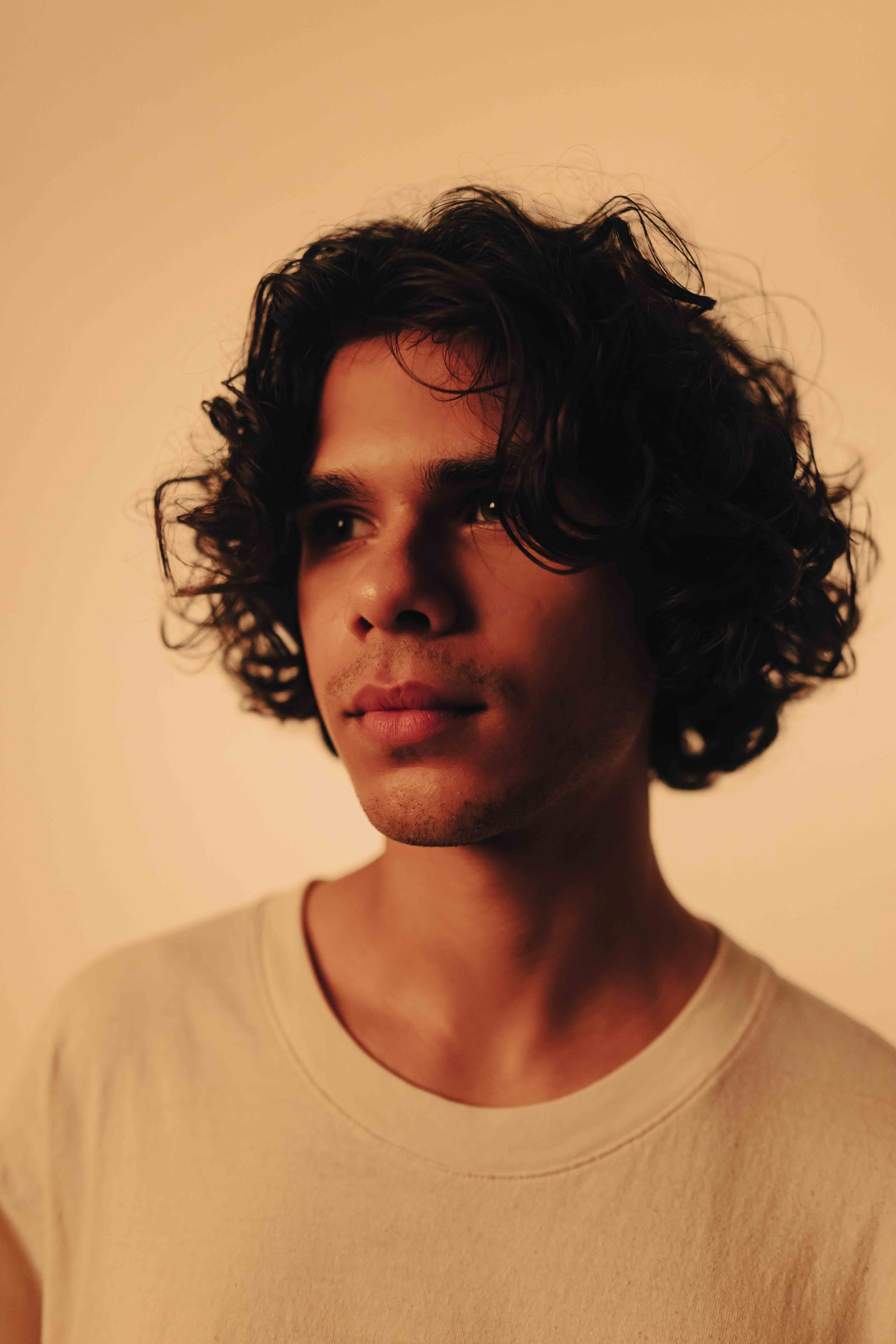 Studio portrait of a young man with curly hair against a warm sand backdrop