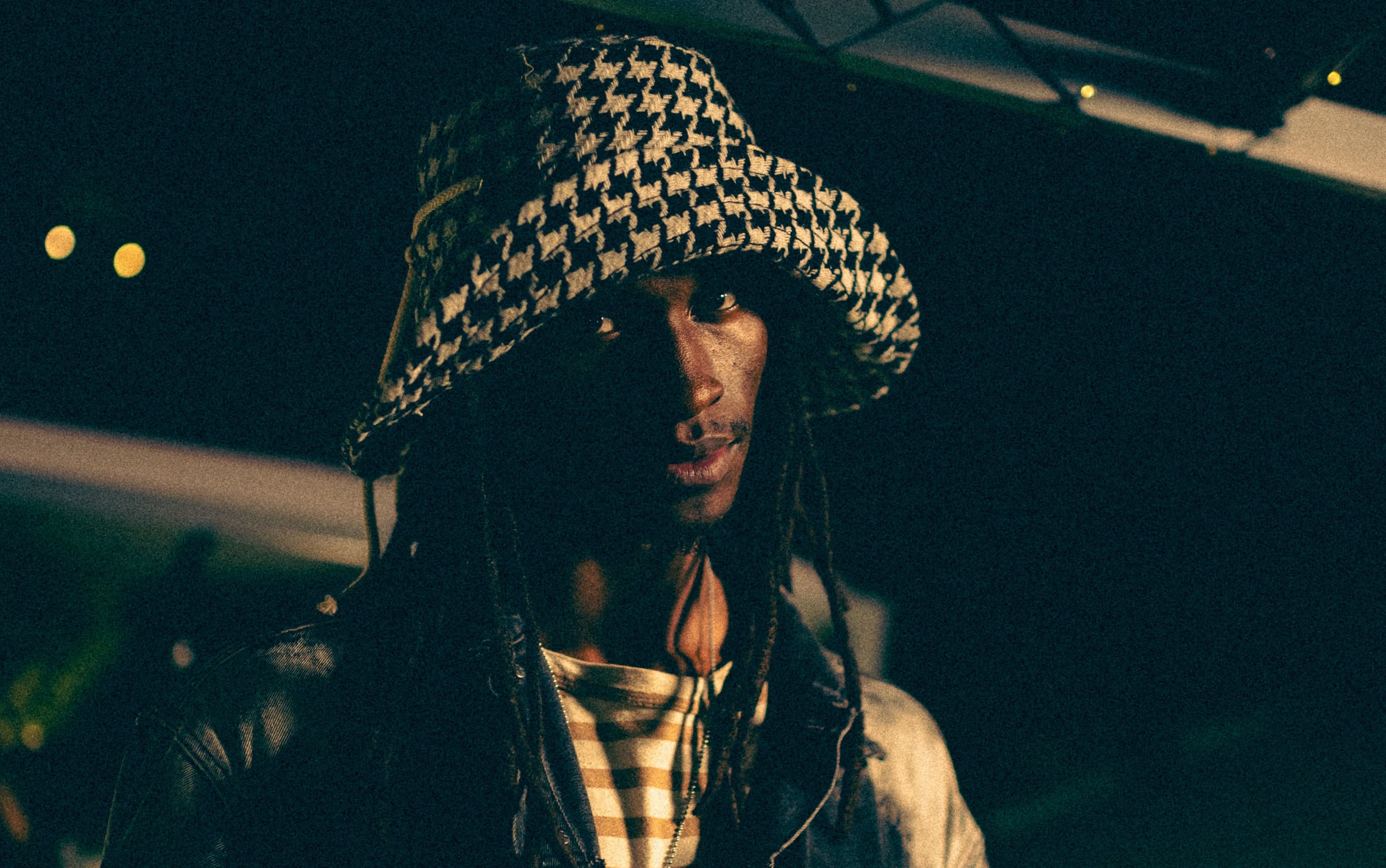 Night portrait of a musician in a houndstooth bucket hat, lit against a dark rooftop