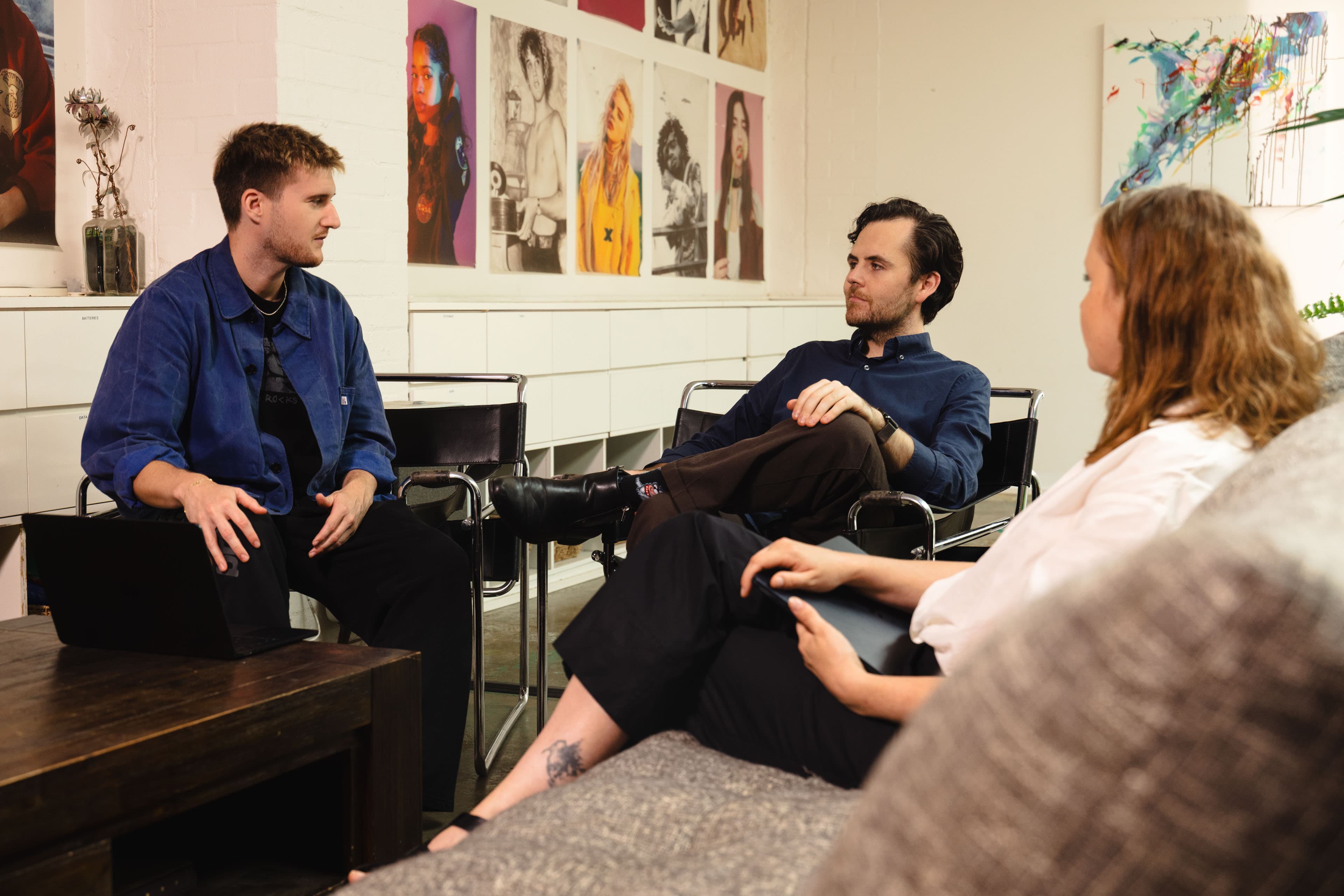 Three people talking in the Mude studio, artwork and prints on the wall behind them